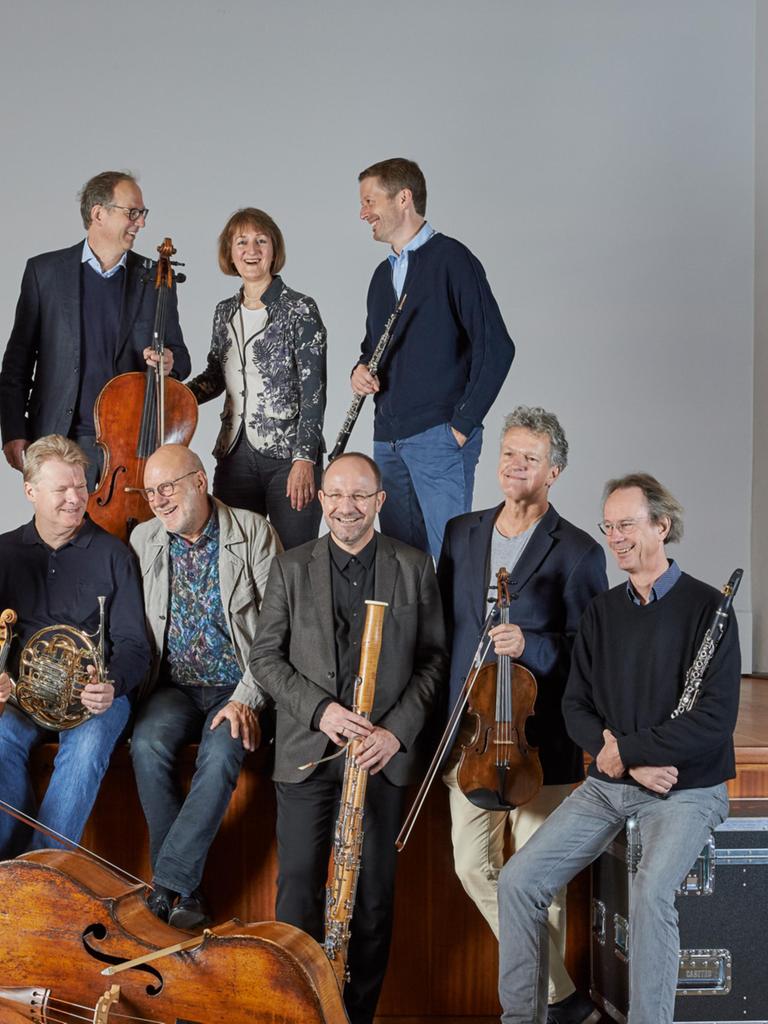 Das Linos Ensemble steht und sitzt mit seinen Instrumenten auf einer Bühne und zum Teil auf Instrumentenkoffern.