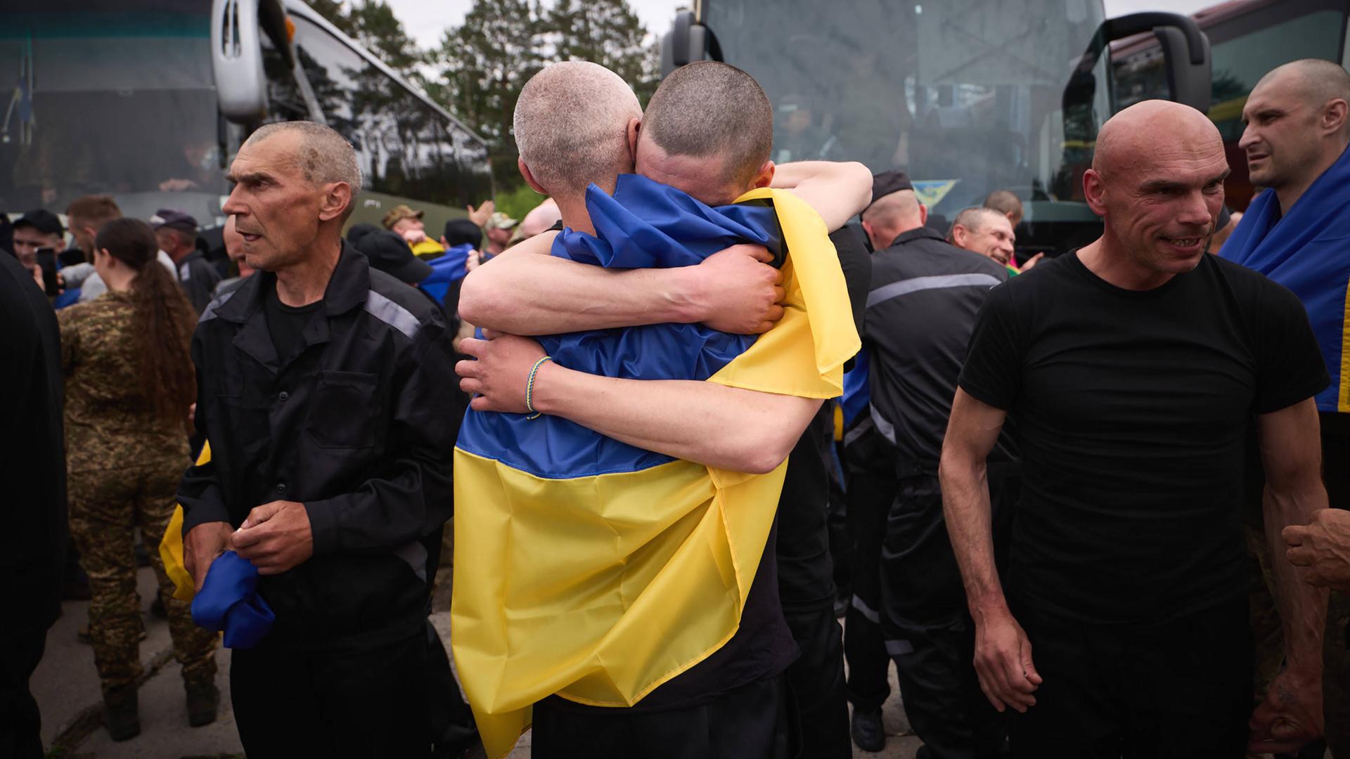 Zwei Männer liegen sich in den Armen, sie sind von einer ukrainischen Flagge bedeckt. Rechts und links von ihnen laufen Männer in schwarzen Klamotten an ihnen vorbei.