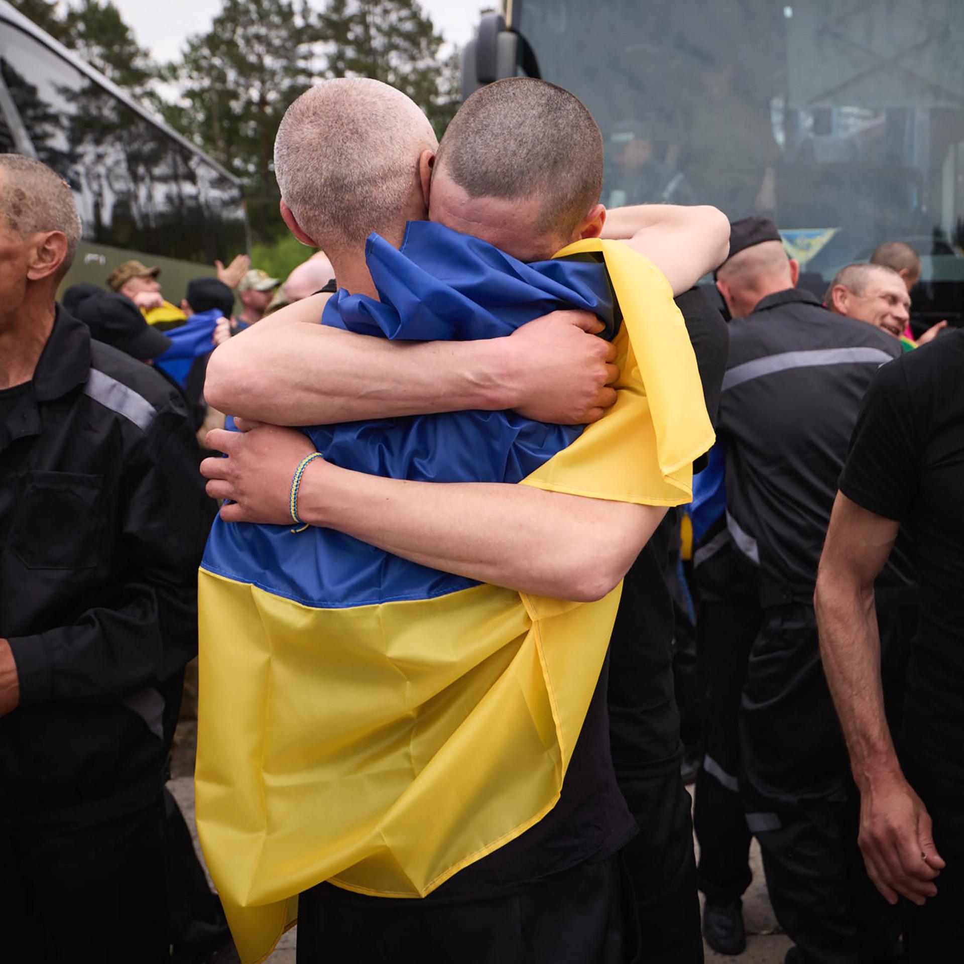 Zwei Männer liegen sich in den Armen, sie sind von einer ukrainischen Flagge bedeckt. Rechts und links von ihnen laufen Männer in schwarzen Klamotten an ihnen vorbei.