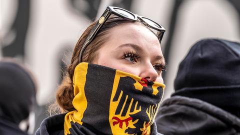 Eine junge Frau auf einer Demo verbirgt mit einem Schal mit dem Bundesadler auf schwarz-rot-goldenem Grund ihre untere Gesichtshälfte.