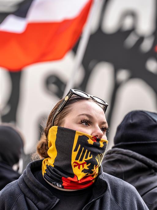 Eine junge Frau auf einer Demo verbirgt mit einem Schal mit dem Bundesadler auf schwarz-rot-goldenem Grund ihre untere Gesichtshälfte.