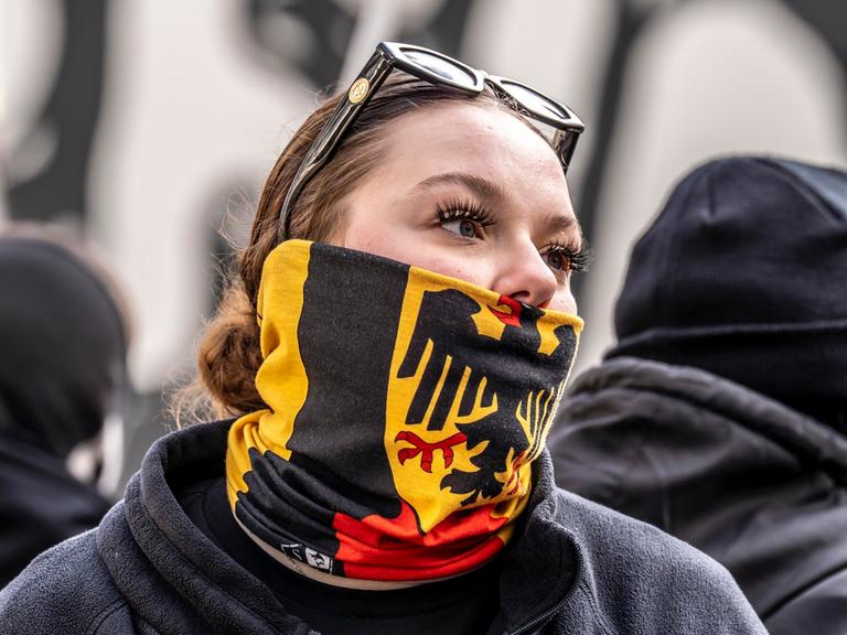 Eine junge Frau auf einer Demo verbirgt mit einem Schal mit dem Bundesadler auf schwarz-rot-goldenem Grund ihre untere Gesichtshälfte.