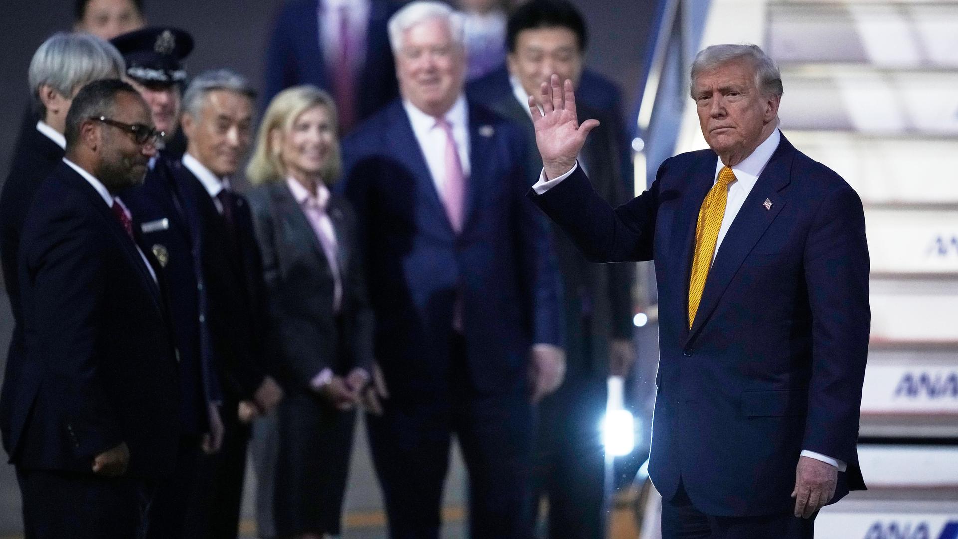 US-Präsident Trump winkt, rechts von ihm stehen verschwommen einige Begleiter.