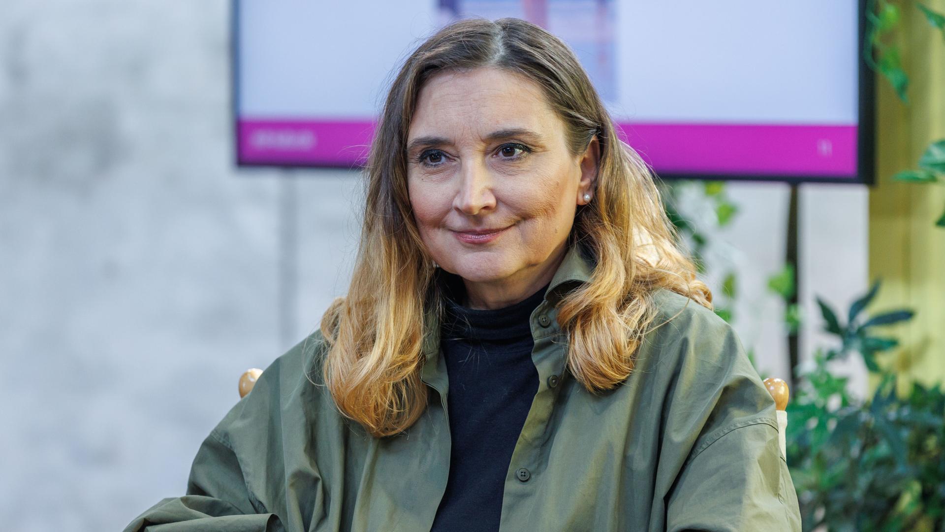 Peggy Patzschke sitzt auf einer Bühne, im Hintergrund ist ein Monitor zu sehen. Sie trägt ein olivgrünes Oberteil. Peggy Patzschke sitzt auf einer Bühne, im Hintergrund ist ein Monitor zu sehen. Sie trägt ein olivgrünes Oberteil.