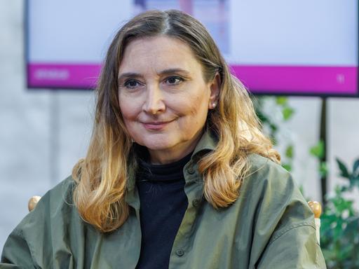 Peggy Patzschke sitzt auf einer Bühne, im Hintergrund ist ein Monitor zu sehen. Sie trägt ein olivgrünes Oberteil. Peggy Patzschke sitzt auf einer Bühne, im Hintergrund ist ein Monitor zu sehen. Sie trägt ein olivgrünes Oberteil.