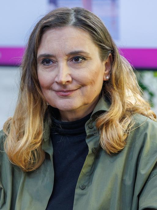 Peggy Patzschke sitzt auf einer Bühne, im Hintergrund ist ein Monitor zu sehen. Sie trägt ein olivgrünes Oberteil.