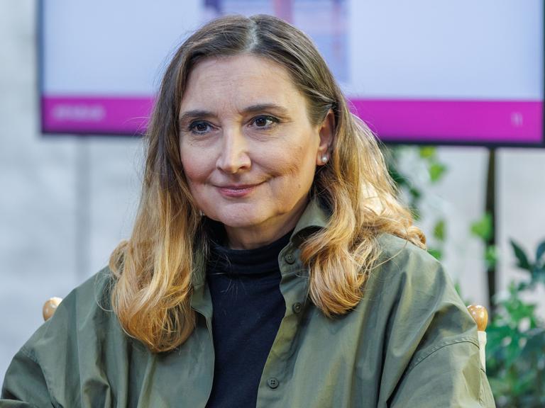 Peggy Patzschke sitzt auf einer Bühne, im Hintergrund ist ein Monitor zu sehen. Sie trägt ein olivgrünes Oberteil.