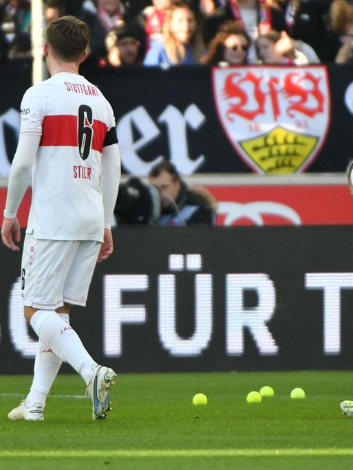 Das Bild zeigt zwei Spieler des VfB in rot-weißen Trikots von hinten. Sie sammeln gelbe Tennisbälle vom Spielfeld auf. Dahinteer unscharf eine Zuschauertribüne. 