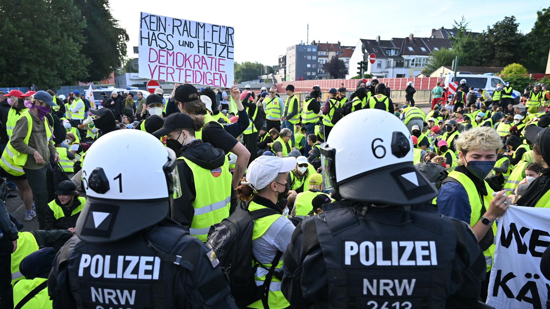 Demonstranten stehen und sitzen auf einer Zufahrtsstraße zum Gelände des AfD-Parteitags in Essen - einige halten ein Schild mit der Aufschrift "Kein Raum für Hass und Hetze - Demokratie verteidigen". Im Bildvordergrund stehen zwei Polizisten.