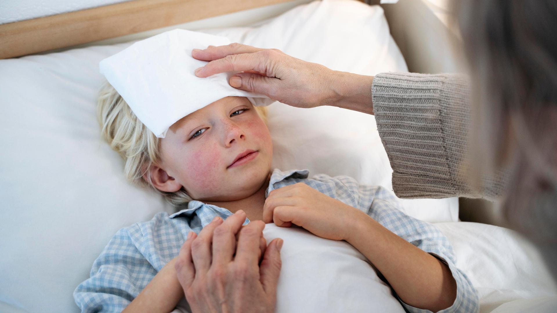 Ein Junge mit Fieber liegt in einem Bett. Eine ältere Frau kühlt ihm mit einem feuchten Tuch die Stirn.