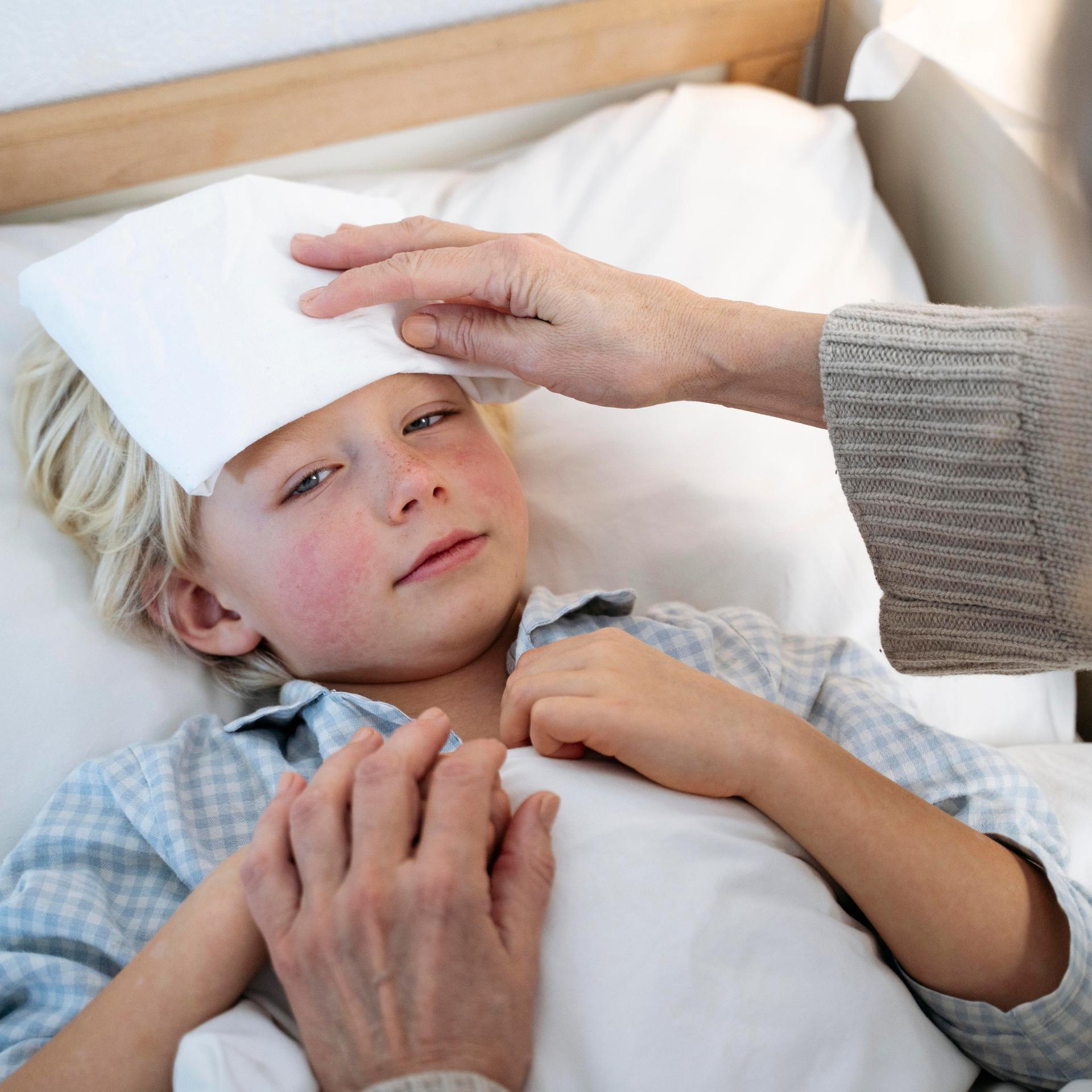 Ein Junge mit Fieber liegt in einem Bett. Eine ältere Frau kühlt ihm mit einem feuchten Tuch die Stirn.