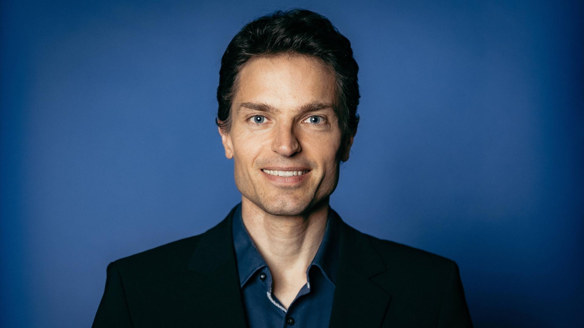 Enzo Weber posiert vor einem blauen Hintergrund für ein Pressefoto.