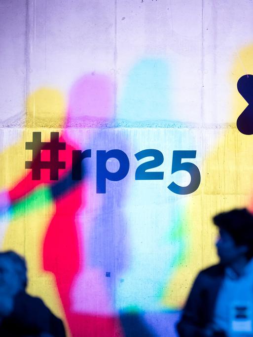 Eine Betonwand wird mit farbigen Projektionen des re:pulica 2025 Logos sowie in Cyan, Gelb und Magenta aufgesplittete Konturen von Menschen bespielt. Im Vordergrund sind die Silhouetten von Besuchern zu erkennen.