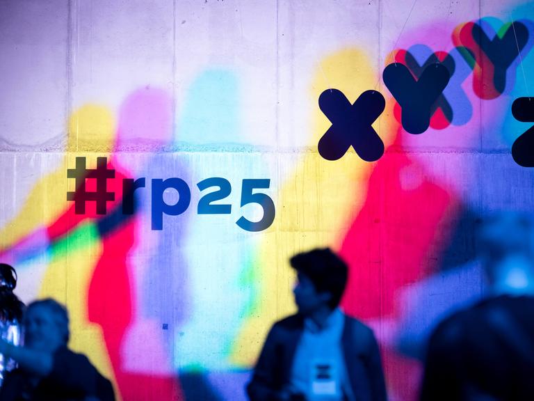 Eine Betonwand wird mit farbigen Projektionen des re:pulica 2025 Logos sowie in Cyan, Gelb und Magenta aufgesplittete Konturen von Menschen bespielt. Im Vordergrund sind die Silhouetten von Besuchern zu erkennen.
