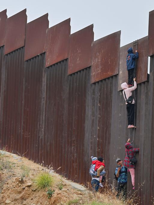 Eine kleine Gruppe von Migranten, darunter mindestens ein Kind, versuchen, mit einer Leiter über die Grenzmauer in die USA zu gelangen.