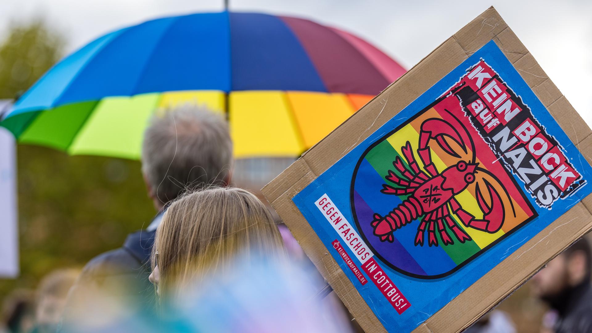 Das Bild zeigt ein Plakat mit der Aufschrift "Kein Bock auf Nazis - Gegen Faschos in Cottbus!"; im Hintergrund ein Schirm mit Regenbogenfarben. Das Bild zeigt ein Plakat mit der Aufschrift "Kein Bock auf Nazis - Gegen Faschos in Cottbus!"; im Hintergrund ein Schirm mit Regenbogenfarben.