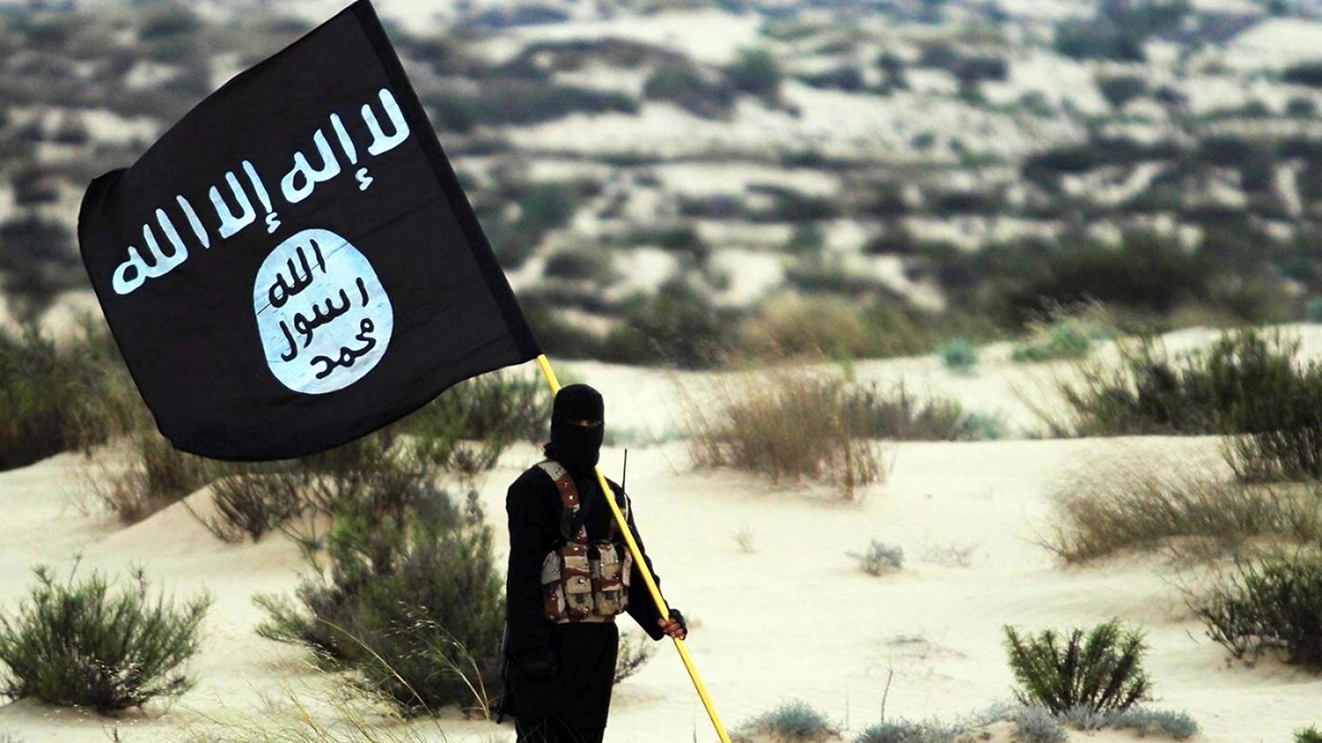 Ein vermummter Terrorist steht mit einer Fahne der Terrororganisation IS in der syrischen Wüste.