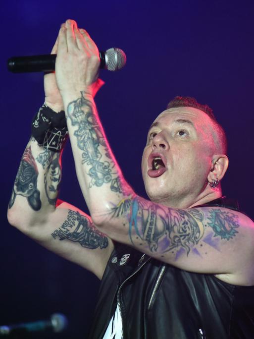 Der Sänger der Punkrockband Tarakany! singt auf einer Bühne. Er hebt seine tätowierten Arme, die das Mikrofon halten.