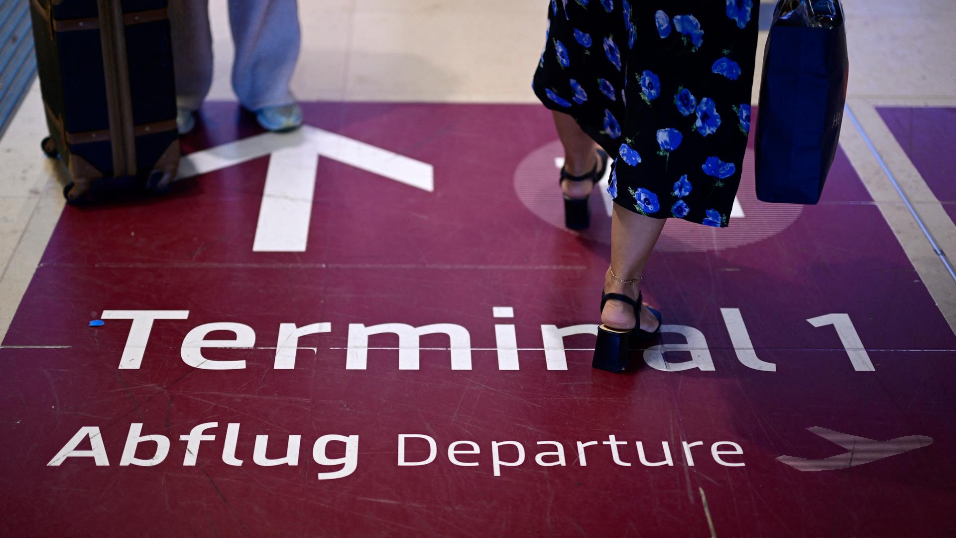 Am Berliner Flughafen BER laufen Menschen durch einen Durchgang. Auf dem Boden befindet sich ein farbig abgesetztes Feld auf dem "Terminal 1, Abflug, Departure" geschrieben steht. Ein Pfeil zeigt die Richtung an. 