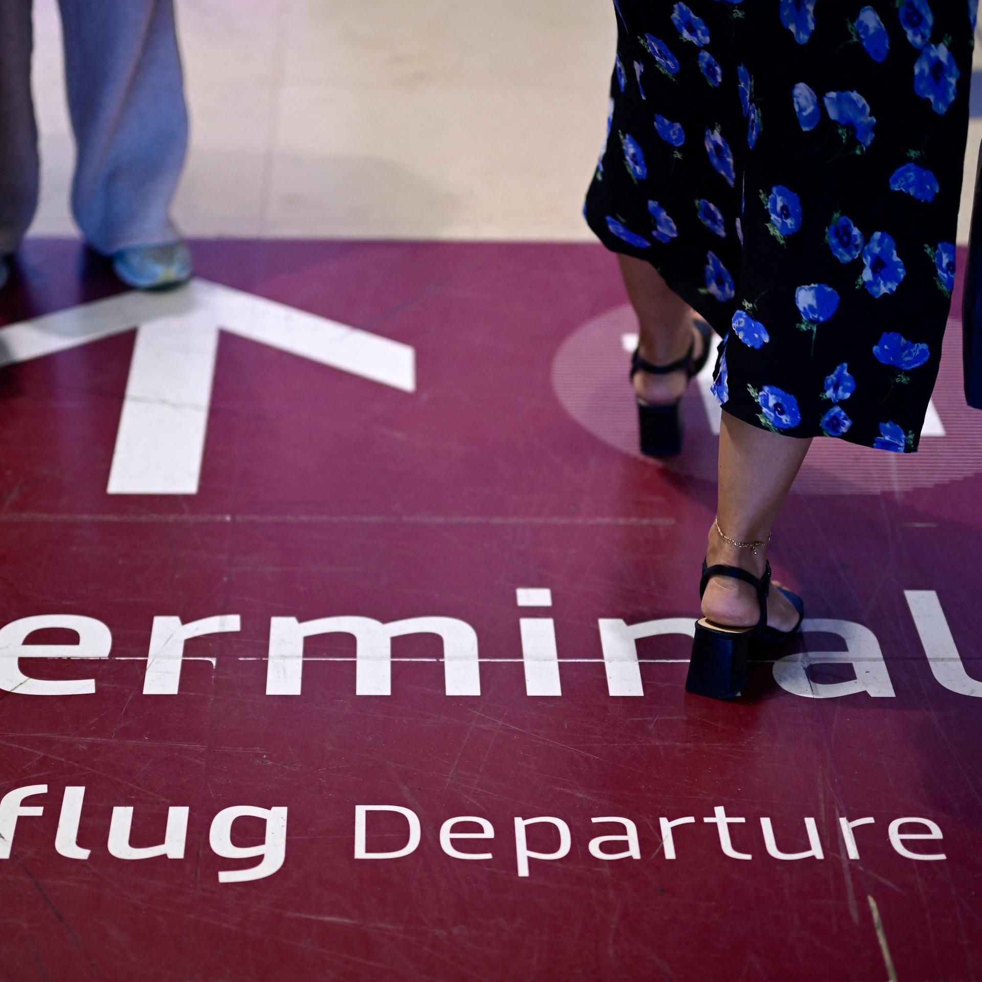 Am Berliner Flughafen BER laufen Menschen durch einen Durchgang. Auf dem Boden befindet sich ein farbig abgesetztes Feld auf dem "Terminal 1, Abflug, Departure" geschrieben steht. Ein Pfeil zeigt die Richtung an. 
