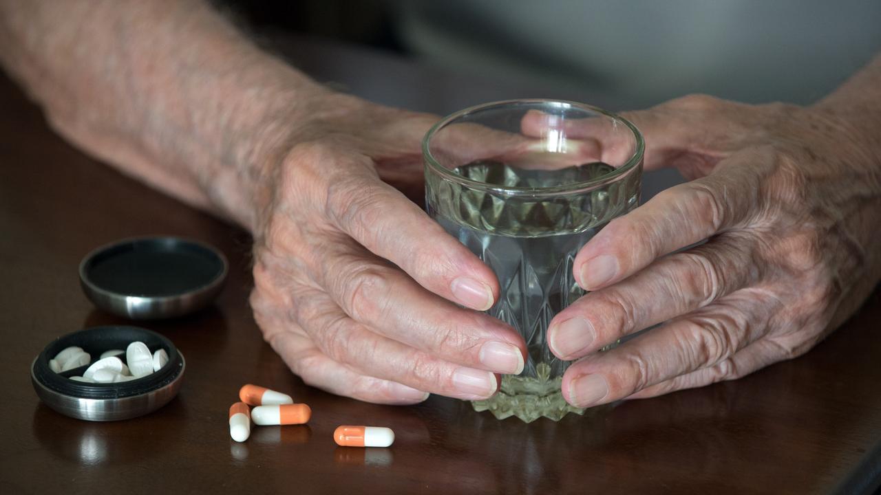Hände eines Senioren umfassen ein gefülltes Wasserglas neben bereit liegenden Tabletten. 