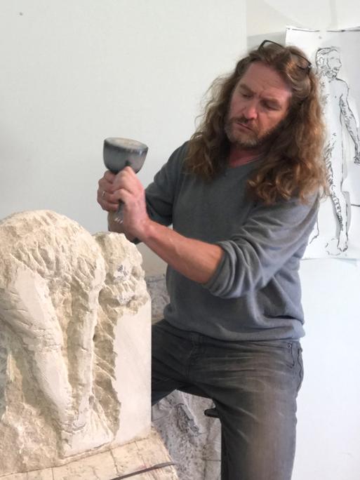 Thomas Lucker arbeitet in seinem Atelier an einer Steinskulptur. Er hat langes braunes Haar und trägt einen grauen Pulli zur Jeans.