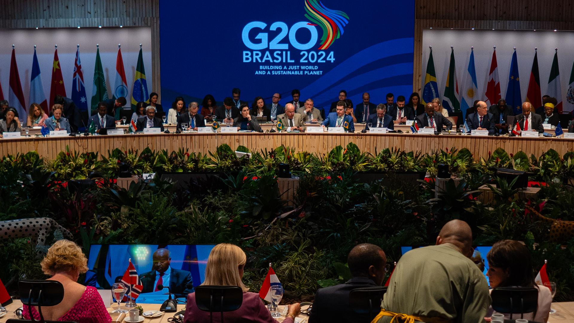 Das Bild zeigt einen Blick in einen großen Konferenz-Saal mit zahlreichen Teilnehmern auf einem Panel und vielen Menschen im Zuschauer-Raum. Über dem Panel hängt ein großes Logo mit der Aufschrift "G20 Brasil 2024"