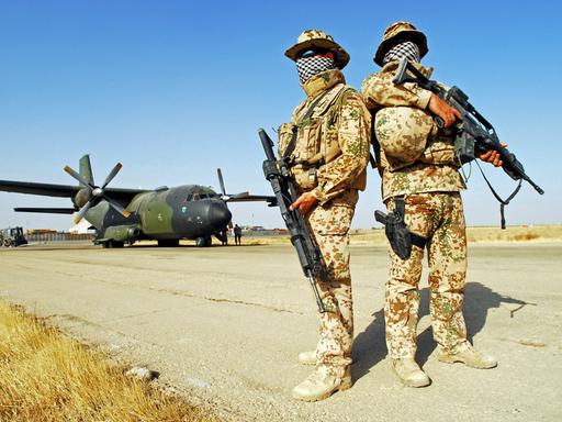 Zwei Soldaten der Bundeswehr stehen vermummt in einer Wüstenlandschaft. Im Hintergrund ein Transportflugzeug.