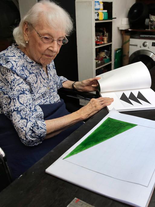 Eine alte Frau sitzt an einem Schreibtisch und blättert durch Zeichungen und Bilder.