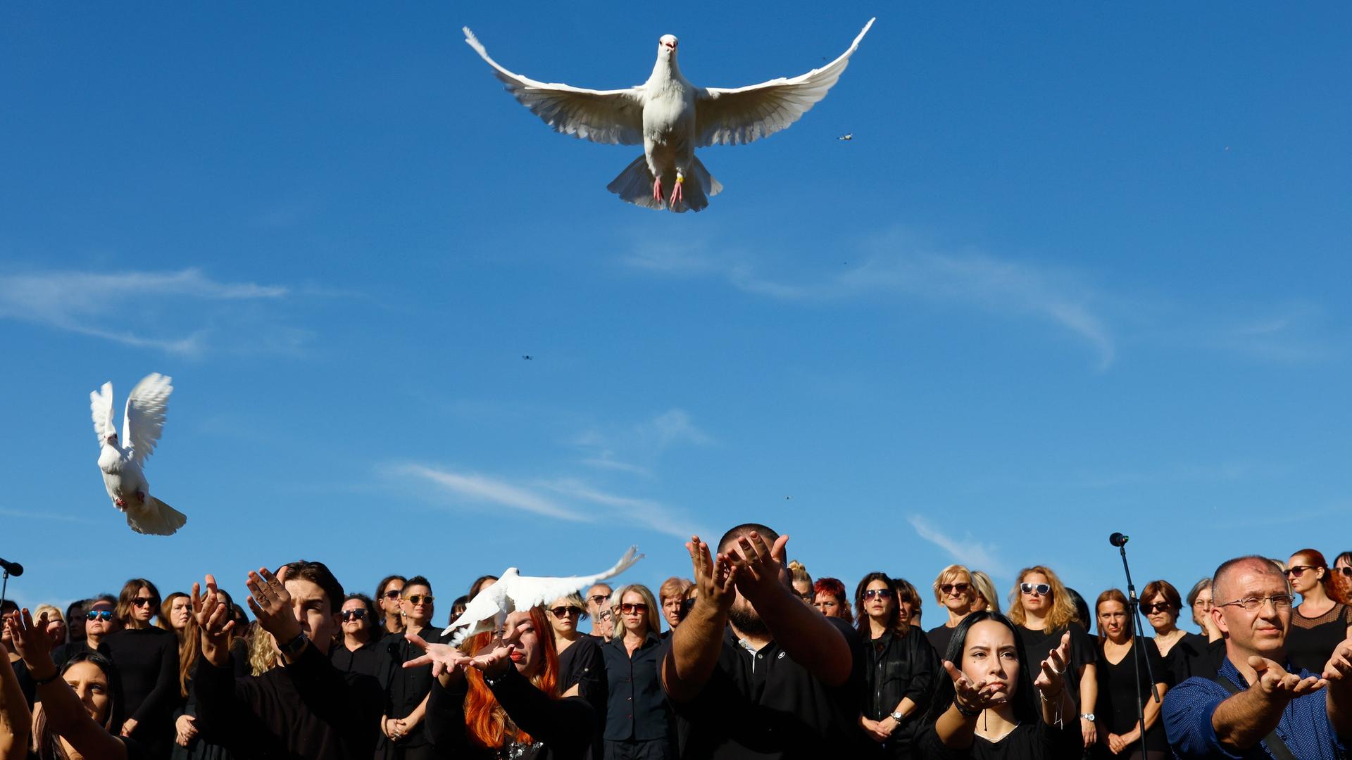 Mehrere Menschen stehen in schwarz gekleidet nebeneinander. Drei von ihnen haben ihre Hände geöffnet und lassen weiße Tauben in den Himmel fliegen. Mehrere Menschen stehen in schwarz gekleidet nebeneinander. Drei von ihnen haben ihre Hände geöffnet und lassen weiße Tauben in den Himmel fliegen.