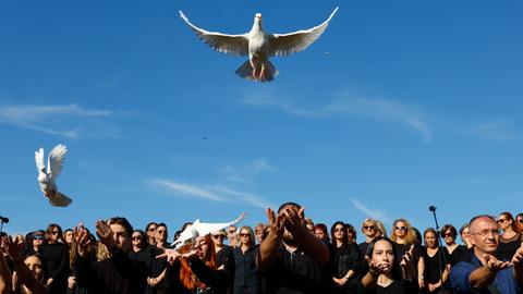 Mehrere Menschen stehen in schwarz gekleidet nebeneinander. Drei von ihnen haben ihre Hände geöffnet und lassen weiße Tauben in den Himmel fliegen.