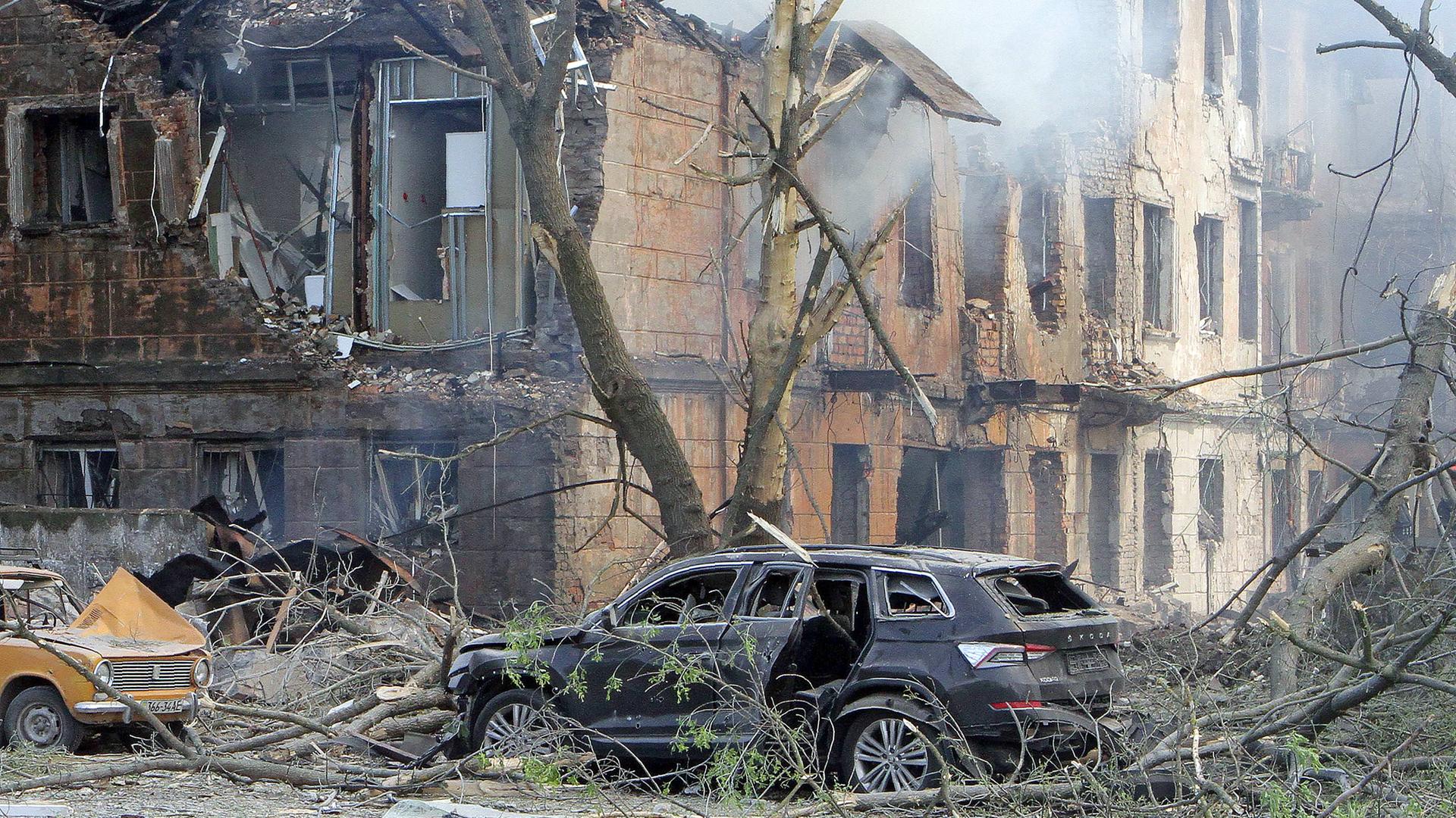 Zwei zerstörte Autos stehen vor einem völlig zerstörten Gebäude in Dnipro. Es steigt Rauch auf. Äste von Bäumen sind abgeknickt.