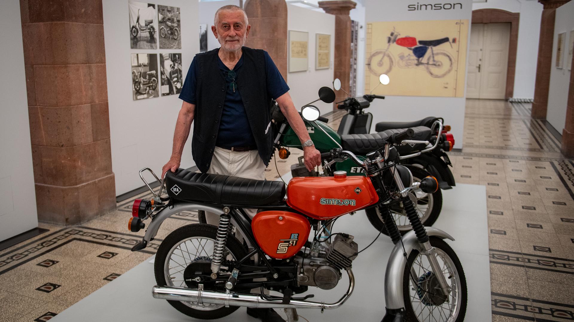 Der Gestalter Karl Clauss Dietel (1934-2022) steht in einer Ausstellung hinter einem Motorrad. 