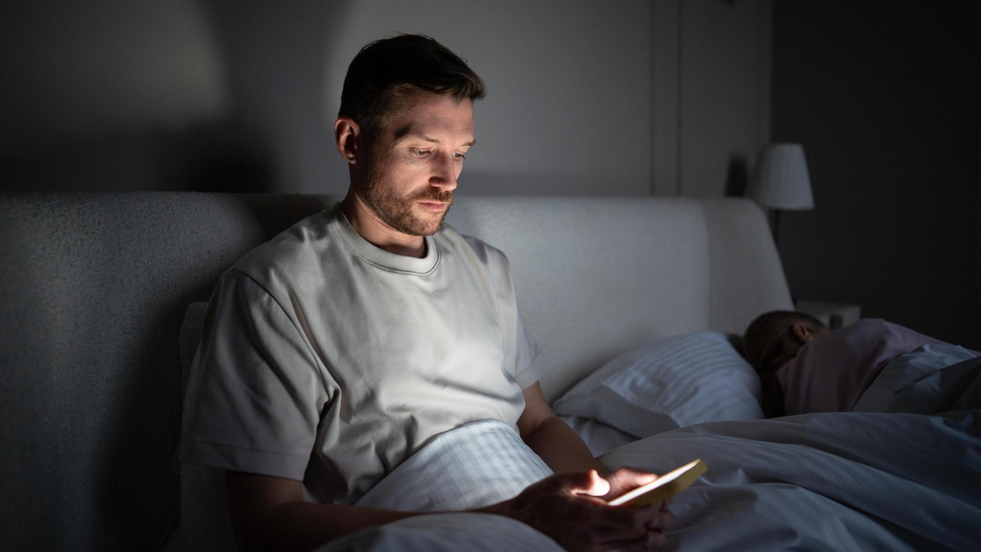 Ein Mann sitzt im Dunkeln auf seinem Bett und starrt in sein Smartphone. Ein Mann sitzt im Dunkeln auf seinem Bett und starrt in sein Smartphone.