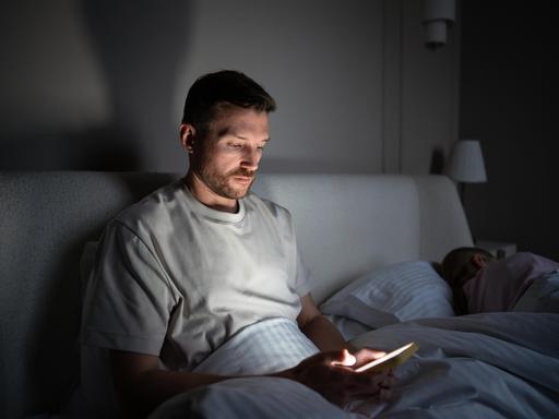 Ein Mann sitzt im Dunkeln auf seinem Bett und starrt in sein Smartphone.