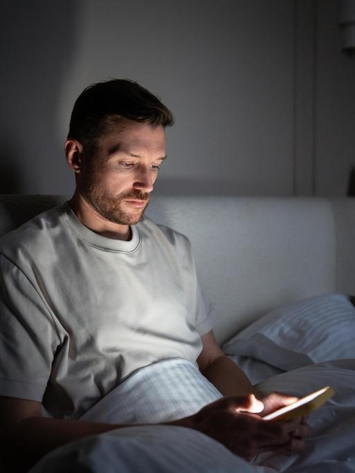 Ein Mann sitzt im Dunkeln auf seinem Bett und starrt in sein Smartphone.