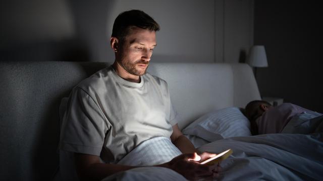 Ein Mann sitzt im Dunkeln auf seinem Bett und starrt in sein Smartphone. Ein Mann sitzt im Dunkeln auf seinem Bett und starrt in sein Smartphone.