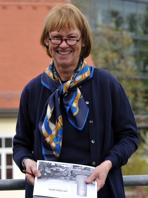 Prof. Dr. Christel Köhle-Hezinger steht vor der Uni Jena, hält ein Buch in der Hand und schaut freundlich in die Kamera. 