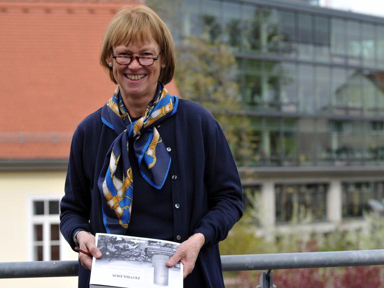 Prof. Dr. Christel Köhle-Hezinger steht vor der Uni Jena, hält ein Buch in der Hand und schaut freundlich in die Kamera. 