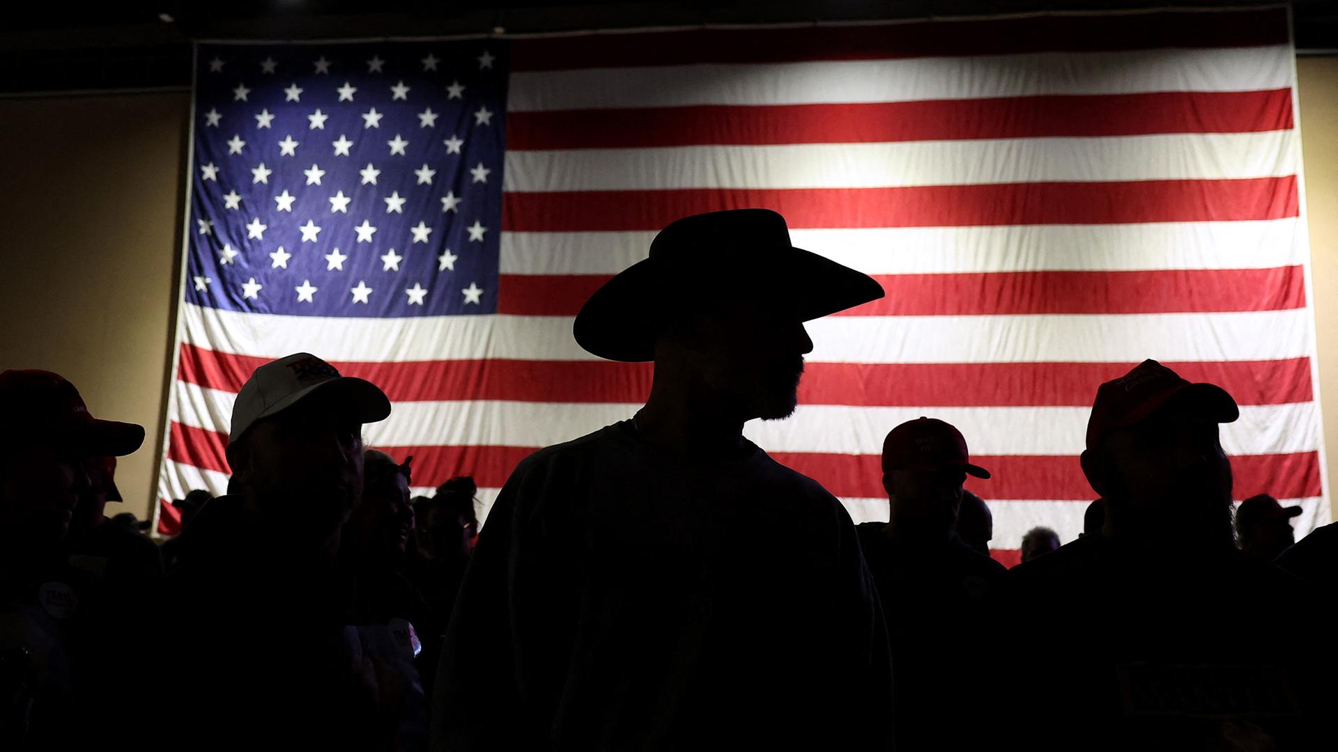 Die Silhouette eines Mannes mit Cowboyhut hebt sich dunkel vor der US-amerikanischen Flagge ab.