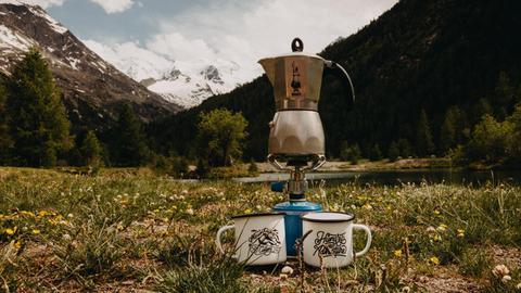 Ein Mokkakocher von Bialetti steht auf einer Bergwiese auf einem Camping-Gaskocher vor einer Alpenszenerie. 