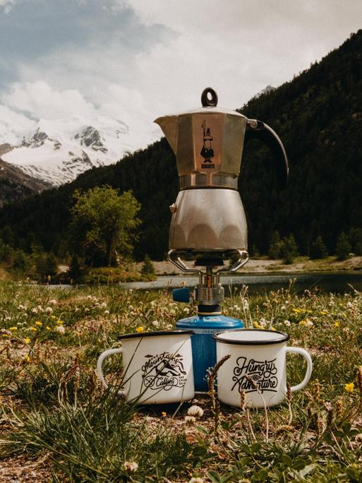 Ein Mokkakocher von Bialetti steht auf einer Bergwiese auf einem Camping-Gaskocher vor einer Alpenszenerie. 