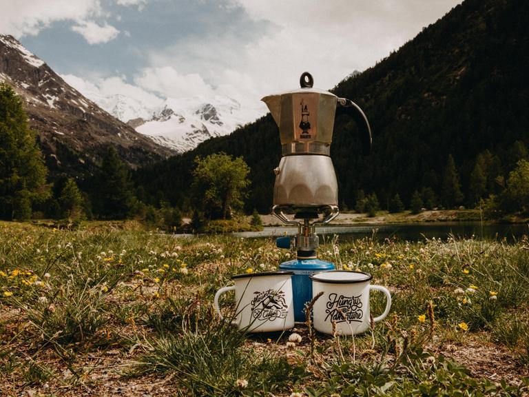 Ein Mokkakocher von Bialetti steht auf einer Bergwiese auf einem Camping-Gaskocher vor einer Alpenszenerie. 