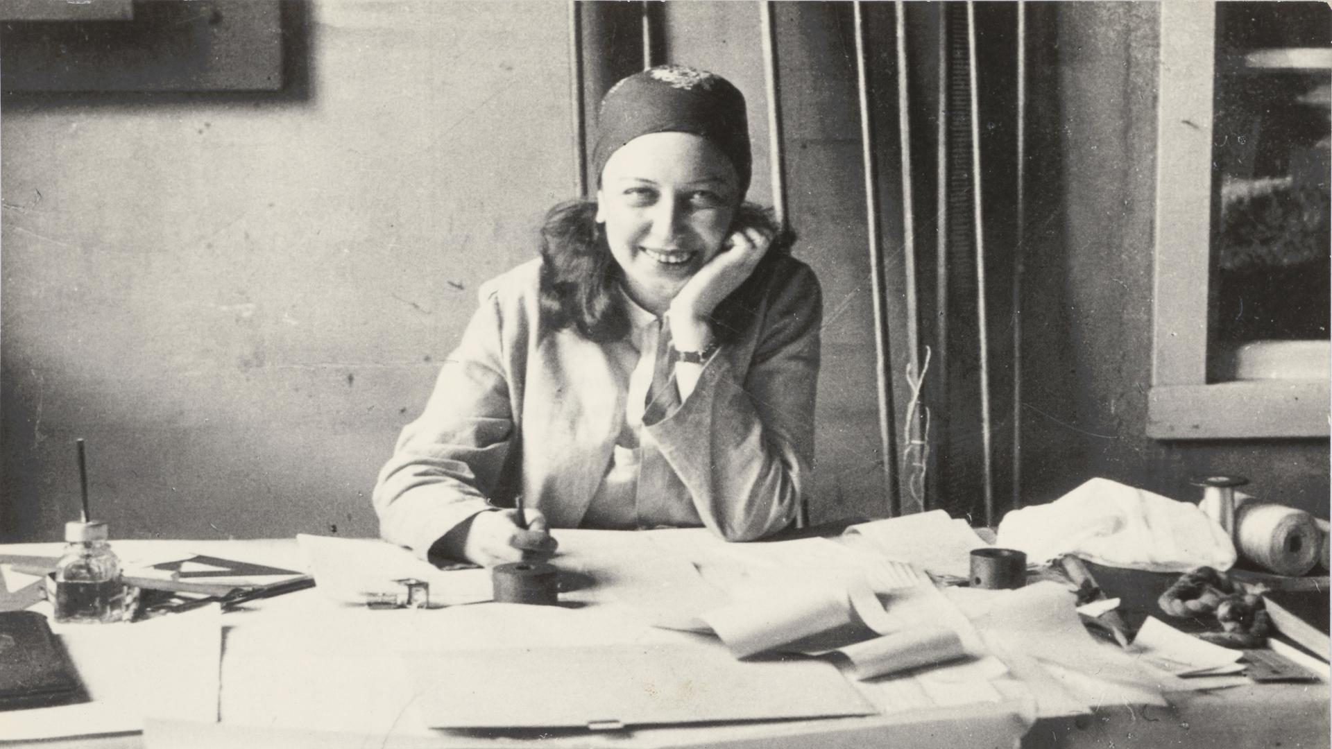 Historisches Schwarzweißfoto von Otti Berger, die lachend an ihrem Arbeitstisch sitzt. Sie trägt einen Blazer und ein gemustertes Tuch um den Kopf gebunden.