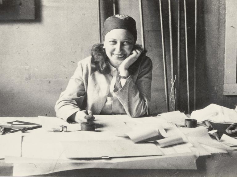 Historisches Schwarzweißfoto von Otti Berger, die lachend an ihrem Arbeitstisch sitzt. Sie trägt einen Blazer und ein gemustertes Tuch um den Kopf gebunden.