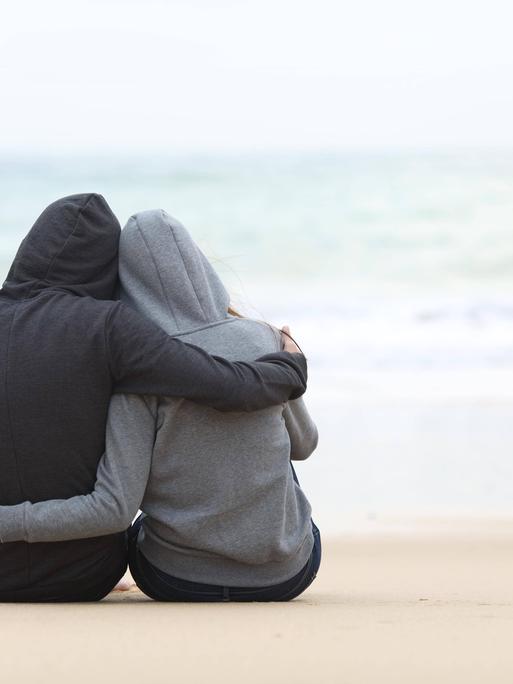 Zwei Menschen in Kapuzenpullis sitzen am Strand und schauen aufs Meer.