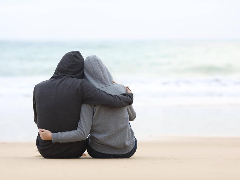 Zwei Menschen in Kapuzenpullis sitzen am Strand und schauen aufs Meer.