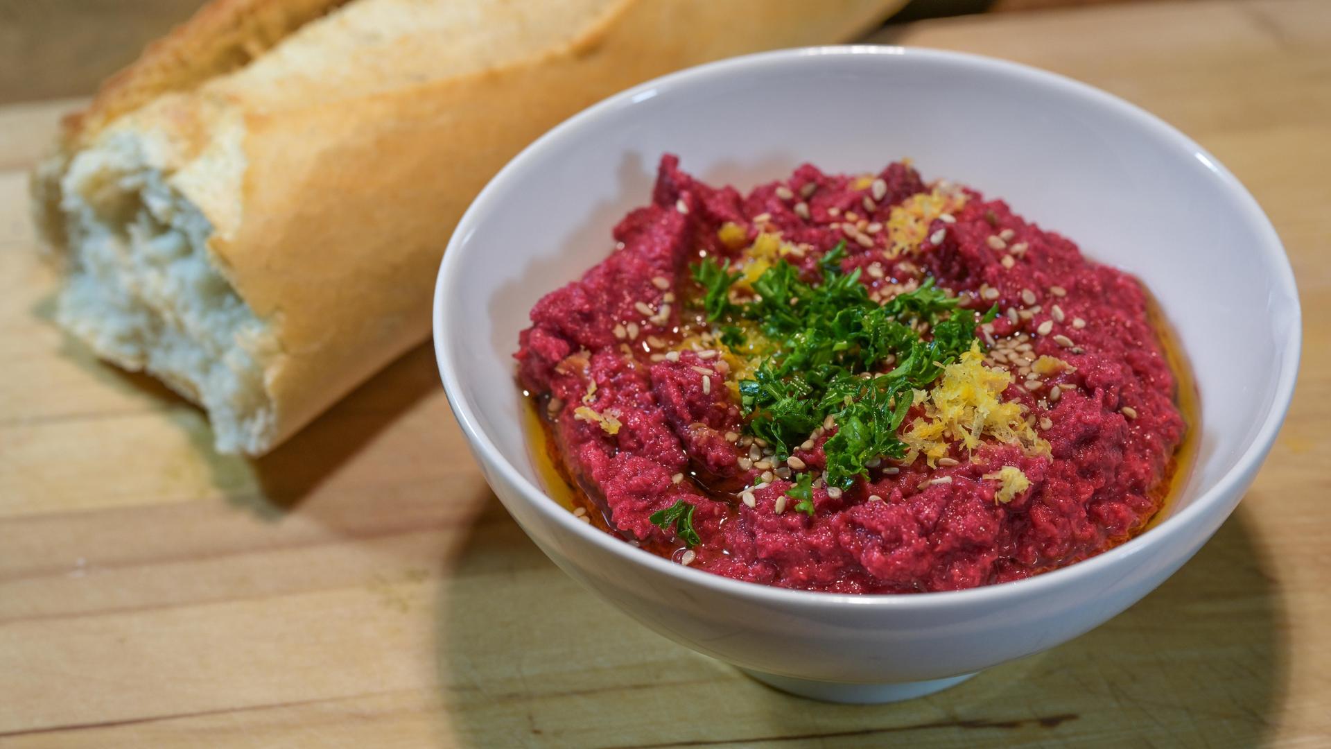 Ausgefallener roter Hummus aus Kichererbsen und Roter Bete mit Sesam, Petersiliengarnitur und Zitronenschale in einer weißen Schale mit Brot auf einem rustikalen Holzbrett, ausgewählter Schwerpunkt