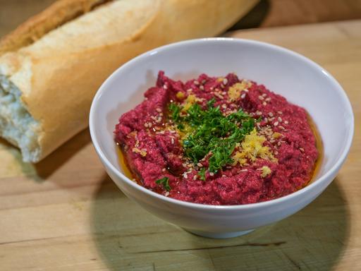Ausgefallener roter Hummus aus Kichererbsen und Roter Bete mit Sesam, Petersiliengarnitur und Zitronenschale in einer weißen Schale mit Brot auf einem rustikalen Holzbrett, ausgewählter Schwerpunkt