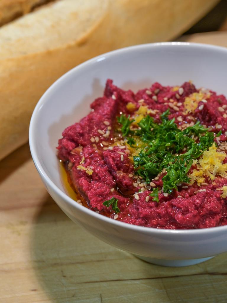 Ausgefallener roter Hummus aus Kichererbsen und Roter Bete mit Sesam, Petersiliengarnitur und Zitronenschale in einer weißen Schale mit Brot auf einem rustikalen Holzbrett, ausgewählter Schwerpunkt